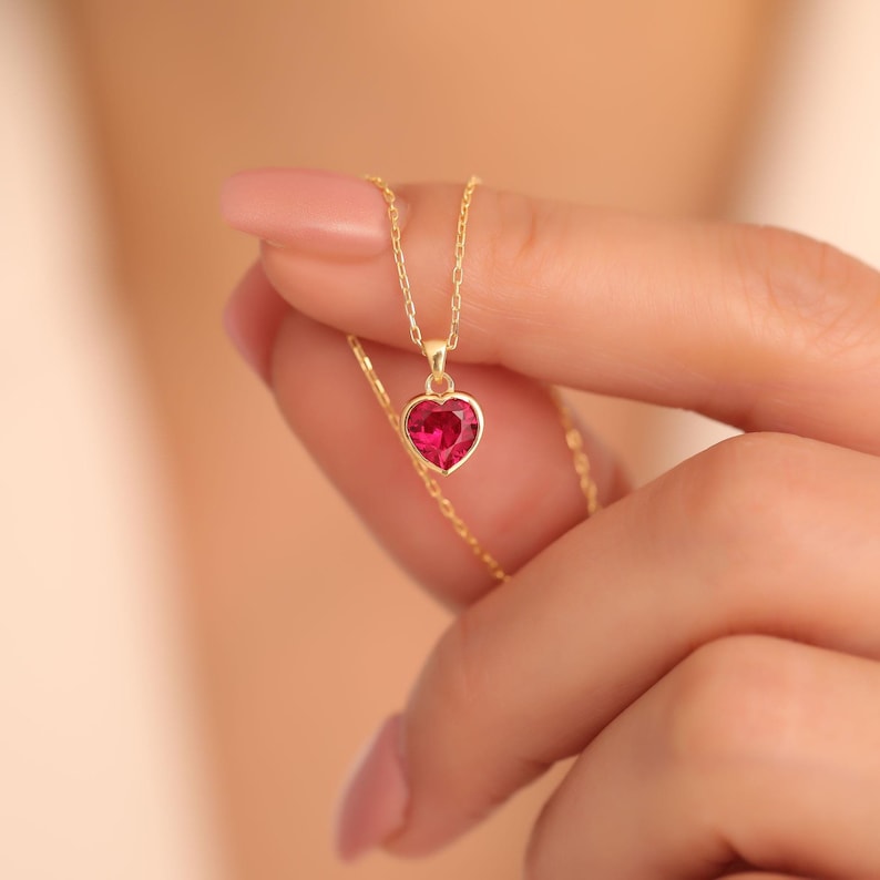 Gold necklace with a heart-shaped pink gemstone held by a hand against a neutral background - gift for her