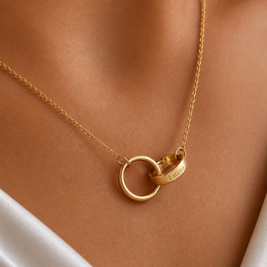A close-up of a gold necklace worn on a model's neck, featuring two interlocking rings with one engraved with the name 'Fatima'.