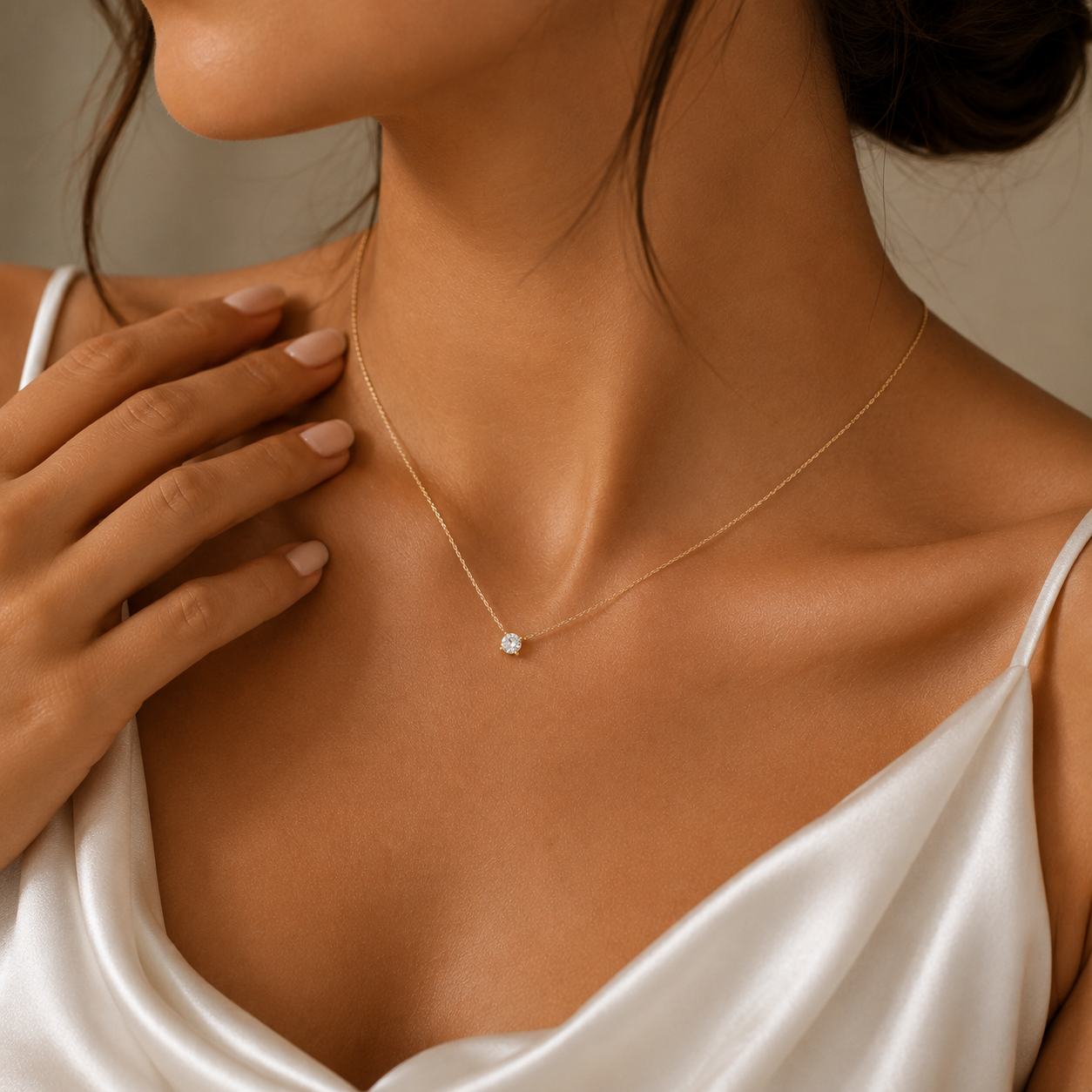 A close-up lifestyle shot of a model wearing a delicate gold chain necklace with a single small clear gemstone pendant, styled with a white satin top.