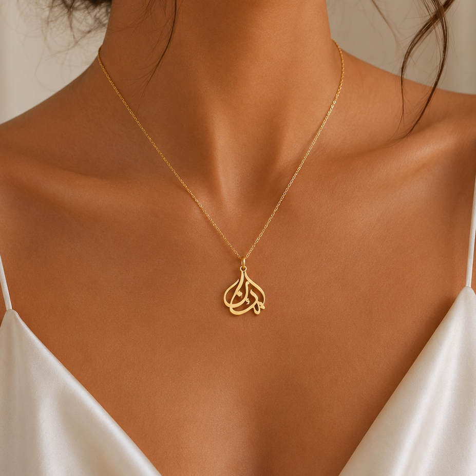 A gold necklace with a stylized interlocking letters in Arabic pendant worn by a model in a white satin camisole top.