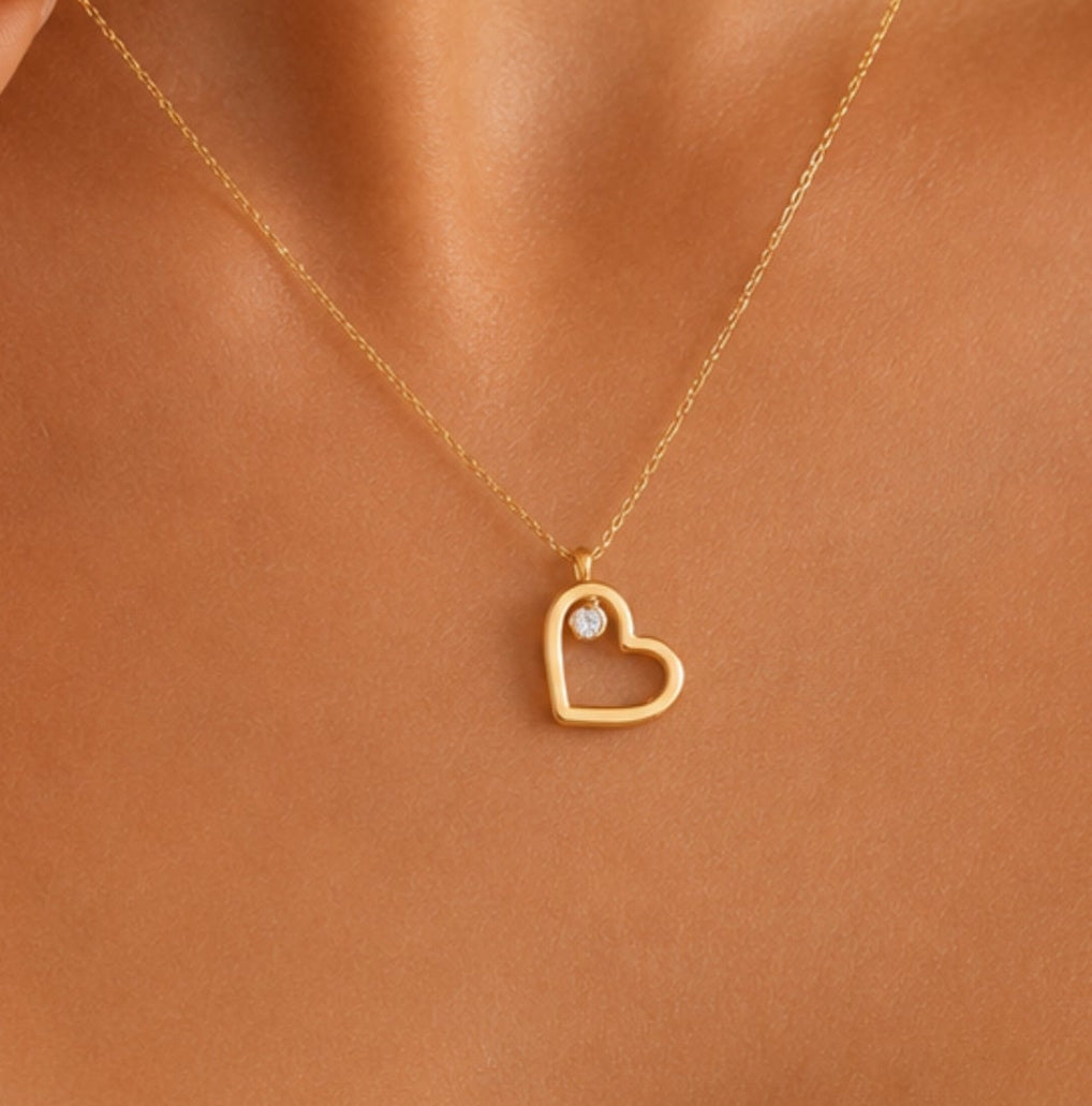A close-up of a model's neck and chest wearing a delicate gold chain necklace with a small open heart pendant and a tiny clear stone.