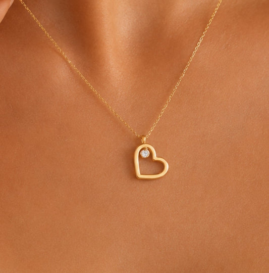 A close-up of a model's neck and chest wearing a delicate gold chain necklace with a small open heart pendant and a tiny clear stone.