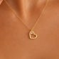 A close-up of a model's neck and chest wearing a delicate gold chain necklace with a small open heart pendant and a tiny clear stone.