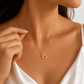 A close-up lifestyle shot of a model wearing a delicate gold chain necklace with a small open heart pendant. The model is wearing a white satin top, and her hand gently touches the chain near the pendant.