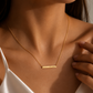 A close-up of a model wearing a delicate gold bar necklace with a date engraved on it, paired with a white satin top.