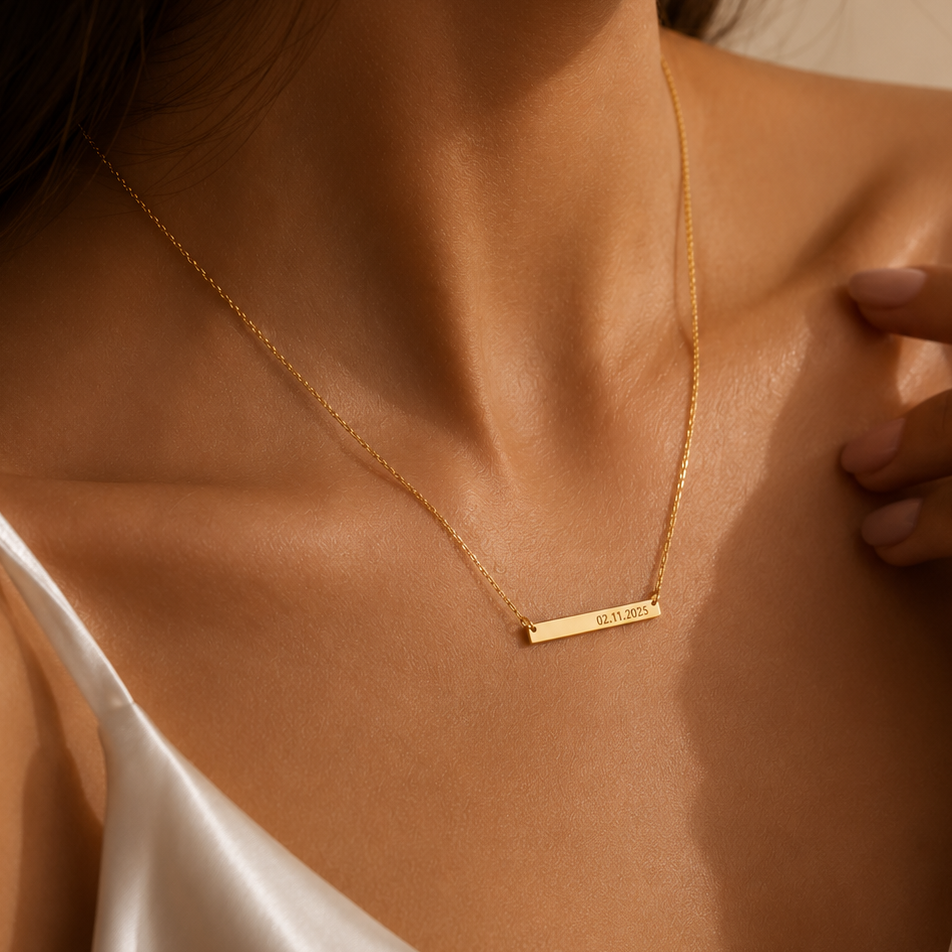 A close-up lifestyle shot of a model wearing a delicate gold bar necklace with a small engraved text, paired with a white satin camisole.