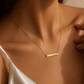 A close-up lifestyle shot of a model wearing a delicate gold bar necklace with a small engraved text, paired with a white satin camisole.
