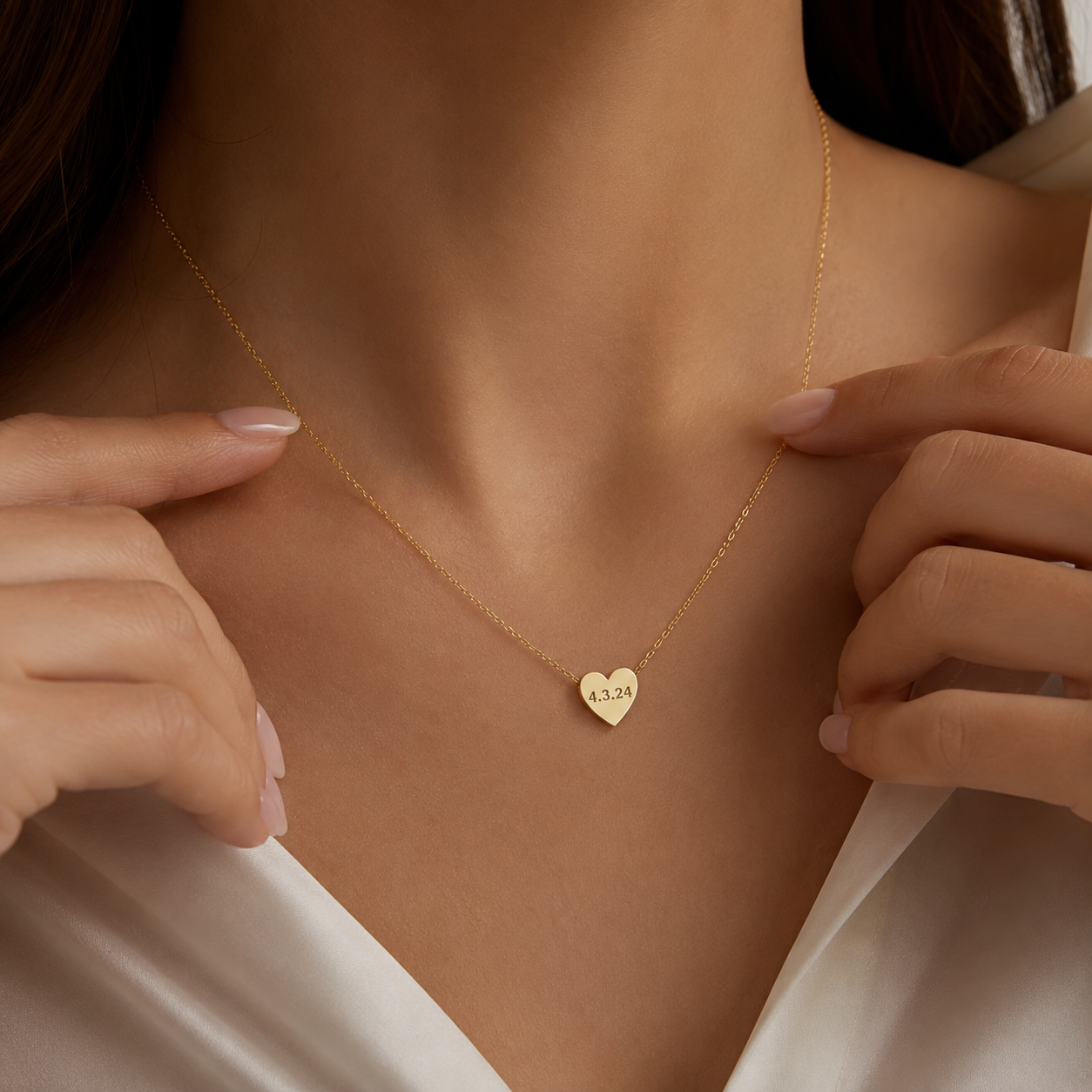 A close-up of a model wearing a delicate gold chain necklace with a small heart-shaped pendant engraved with the numbers '12-23'. The model is wearing a white satin top, and her hands are gently touching the necklace.