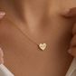 A close-up of a delicate gold chain necklace worn on a model's neck, featuring a small heart-shaped pendant engraved with the date '4.3.