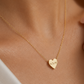 A close-up of a model wearing a delicate gold chain necklace with a small engraved gold heart pendant. The pendant features the initials 'E.