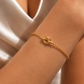 A delicate gold chain bracelet with a knotted center detail, shown worn on a person's wrist against a soft champagne satin background.