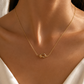 A delicate gold chain necklace with a small interlocking knot pendant, worn on a model's neck. The model is wearing a white satin top, and the background is a soft, warm-toned neutral wall with natural light shadows.