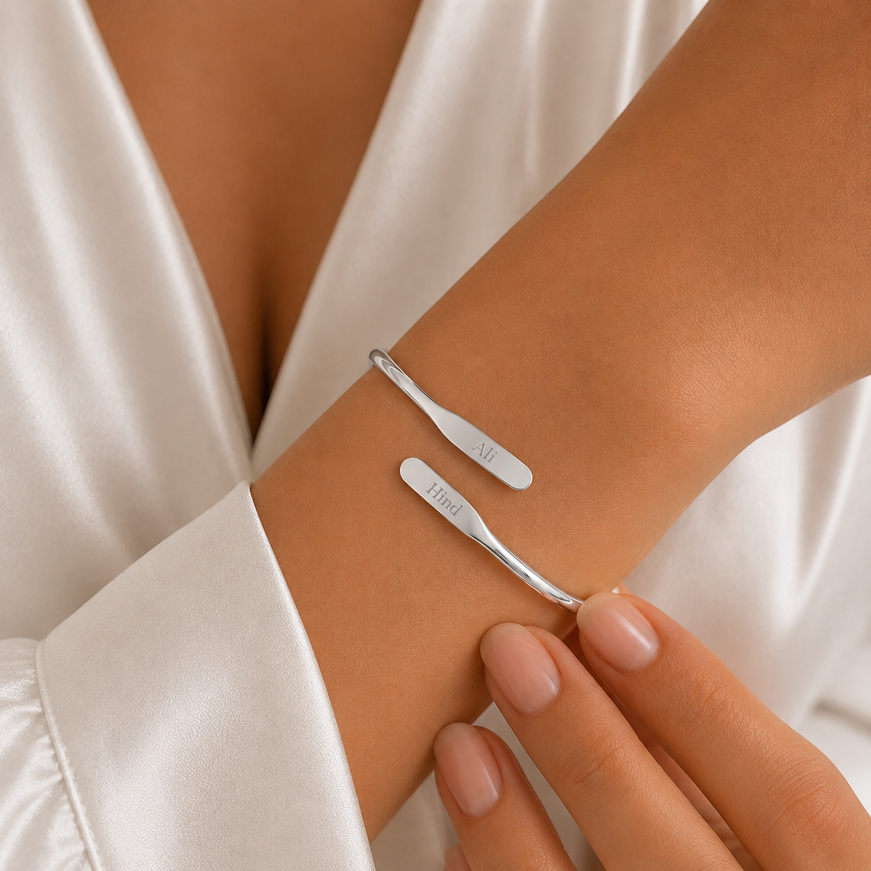 A close-up of a person's wrist wearing two slim silver cuff bracelets, one engraved with the names Ali and Hind, against a white satin robe.