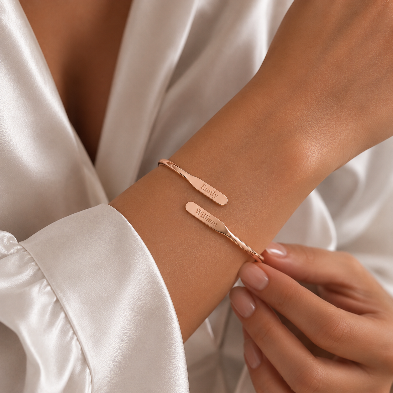 A close-up lifestyle shot of a person wearing a delicate rose gold cuff bracelet with custom engraved names, 'EMILY' and 'WILLIAM', on a satin white robe.