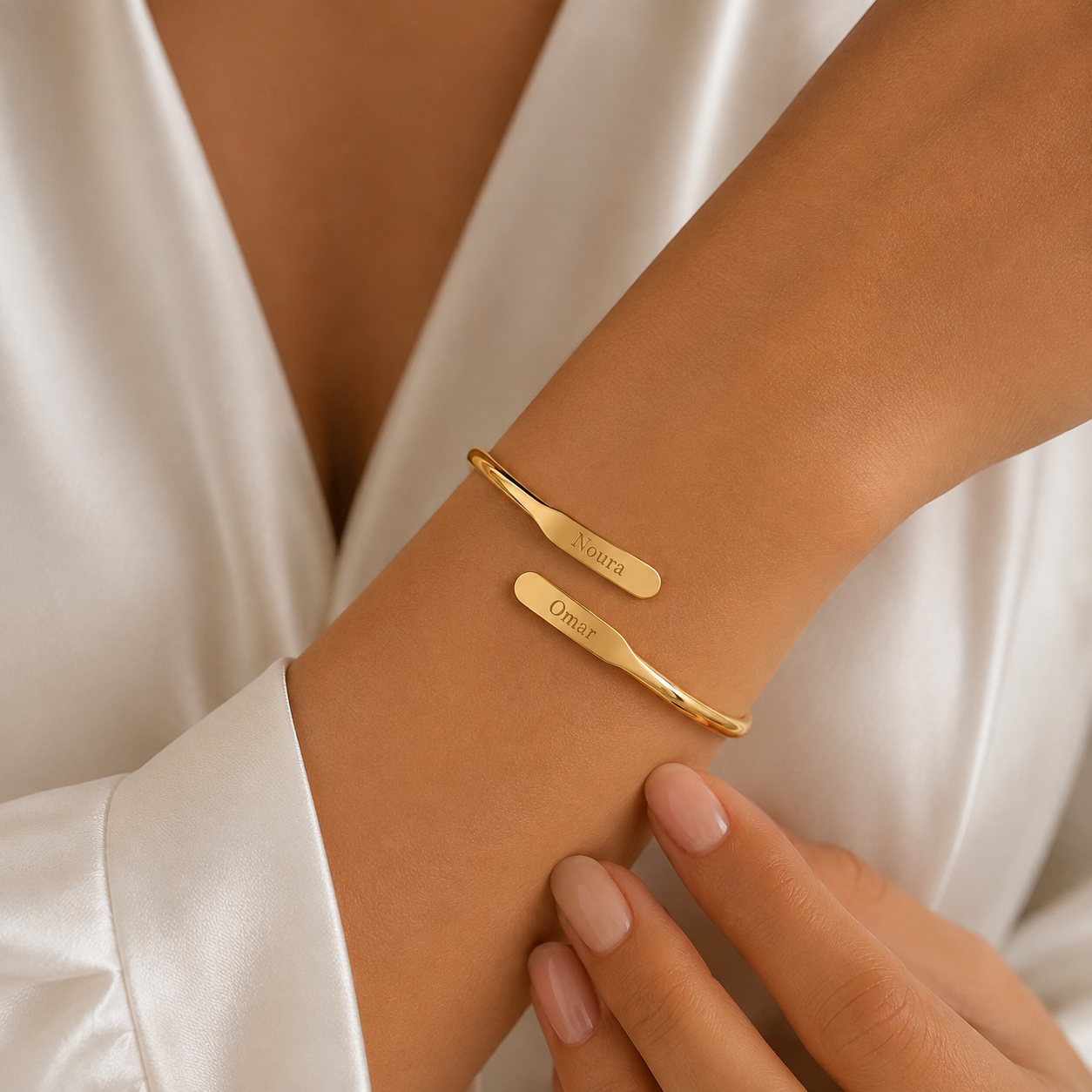 A close-up of a person's wrist wearing two slim gold cuff bracelets, one engraved with the name 'Noura' and the other with 'Omar'. The person is wearing a white satin robe, and their hand with manicured nails is resting near the bracelets.