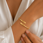 A close-up of a person's wrist wearing two slim gold cuff bracelets, one engraved with the name 'Noura' and the other with 'Omar'. The person is wearing a white satin robe, and their hand with manicured nails is resting near the bracelets.