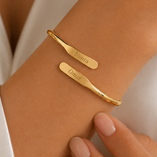 Two slim gold cuff bracelets engraved with the names 'Noura' and 'Omar', shown worn on a person's wrist with fingers visible.
