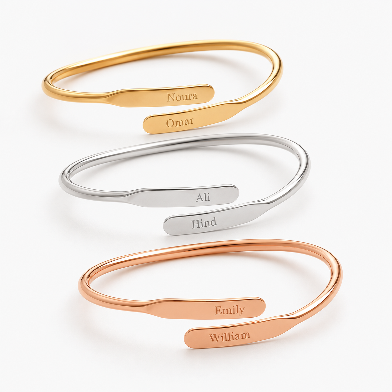 Four minimalist open cuff bracelets arranged vertically on a white background, each in a different metal finish: gold, silver, and rose gold. Each bracelet has a flat engraved nameplate with a personalized name: Noura, Omar, Ali, and Emily for the gold and rose gold, and Hind, William for the silver.