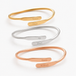 Four minimalist open cuff bracelets arranged vertically on a white background, each in a different metal finish: gold, silver, and rose gold. Each bracelet has a flat engraved nameplate with a personalized name: Noura, Omar, Ali, and Emily for the gold and rose gold, and Hind, William for the silver.