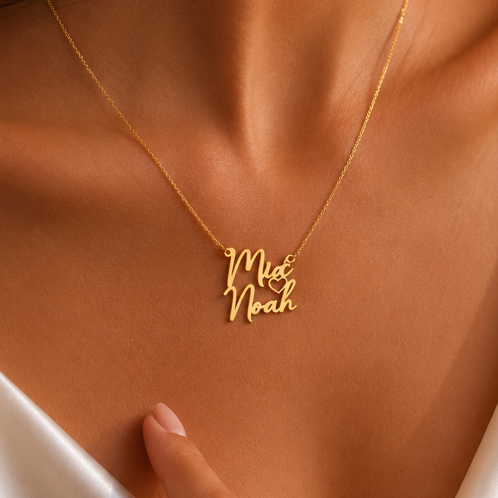 A close-up lifestyle shot of a woman wearing a delicate gold necklace with a script pendant that reads 'Mia Noah'. She is wearing a white satin camisole against a warm, neutral background.