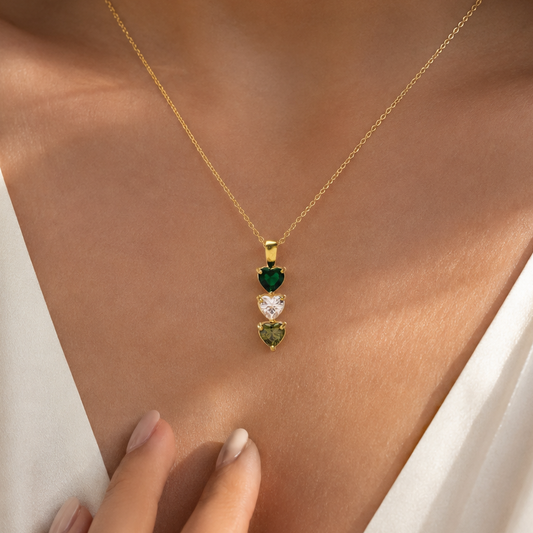 A close-up of a woman wearing a delicate gold chain necklace with three small heart-shaped gemstones in green, white, and green, styled with a white satin top.