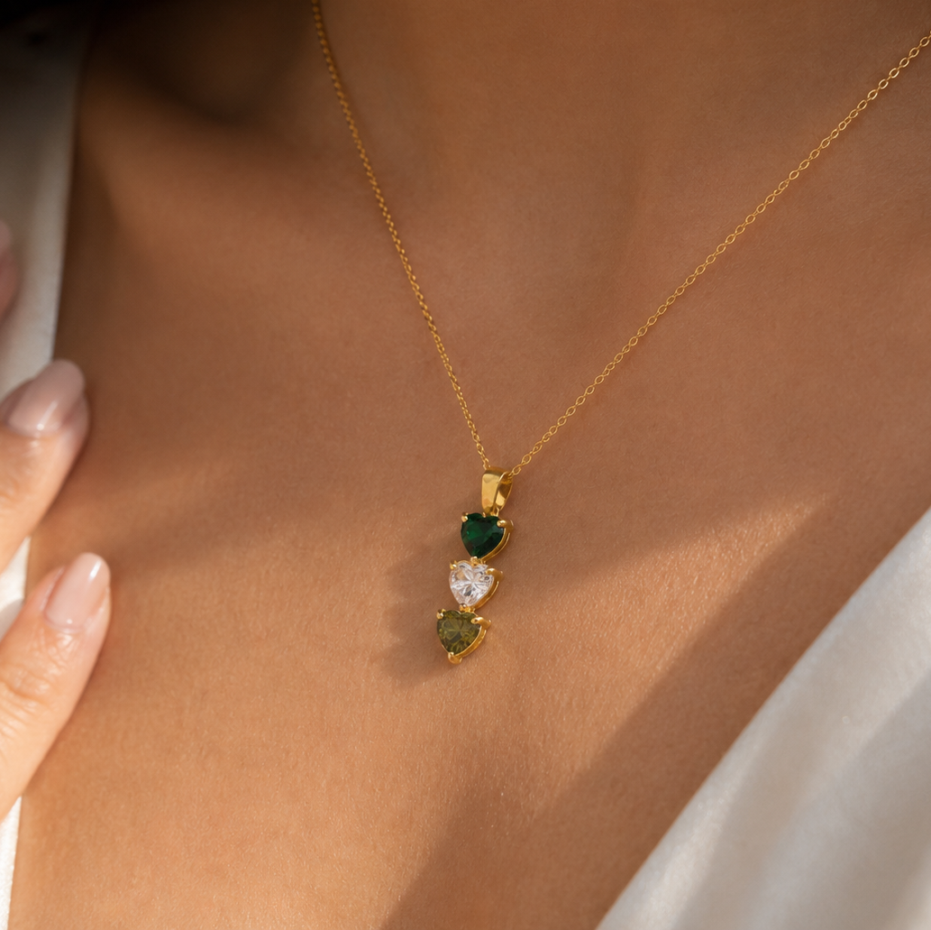 A close-up of a model wearing a delicate gold necklace with three small heart-shaped gemstones in green and clear, styled with a white top.