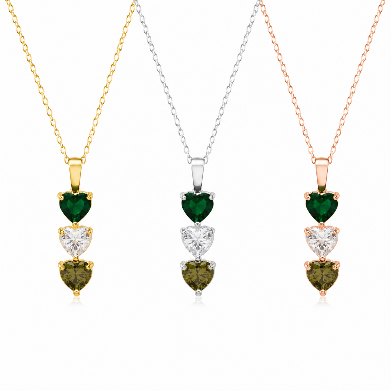 Three heart-shaped pendant necklaces on delicate chains, each featuring stacked heart gemstones: a green emerald, a clear round stone, and a green marquise stone. Shown in yellow gold, silver, and rose gold settings against a white background.