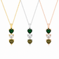 Three heart-shaped pendant necklaces on delicate chains, each featuring stacked heart gemstones: a green emerald, a clear round stone, and a green marquise stone. Shown in yellow gold, silver, and rose gold settings against a white background.
