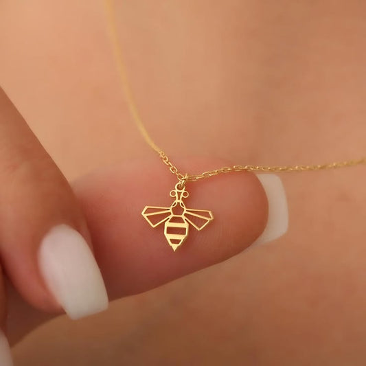 A delicate gold bee pendant necklace held on a person's finger against a warm skin-toned background.