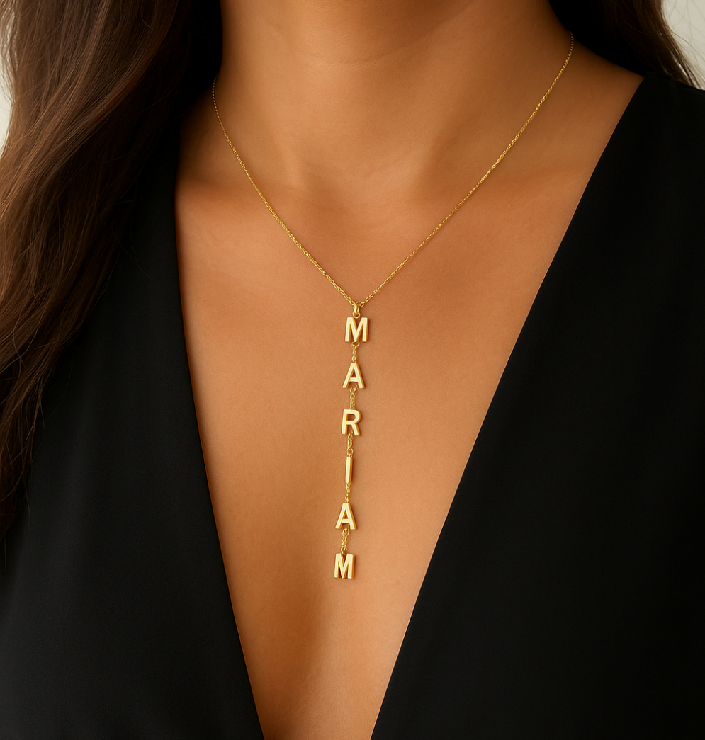 Gold Y-shaped lariat necklace with name "MARIAM" in capital letter charms, worn by woman in black V-neck dress. Personalized gold jewelry for women.