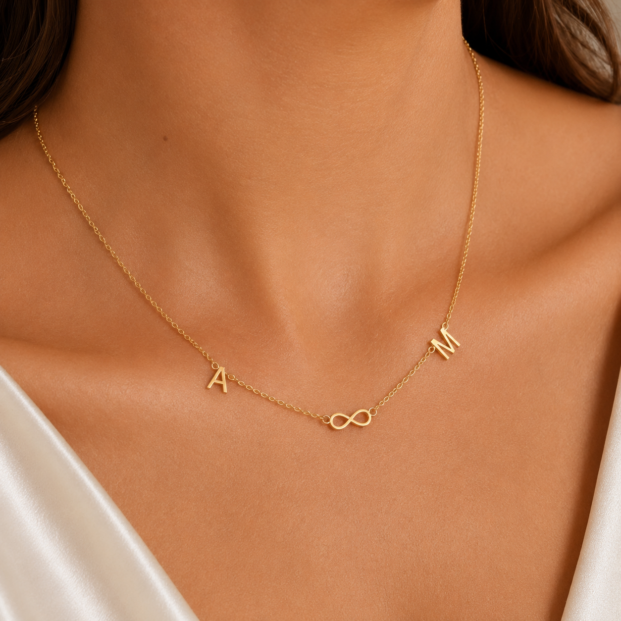 A delicate gold chain necklace worn on a model's neck, featuring the letters 'A' and 'M' at the ends and a small infinity symbol in the center, styled with a white satin top.