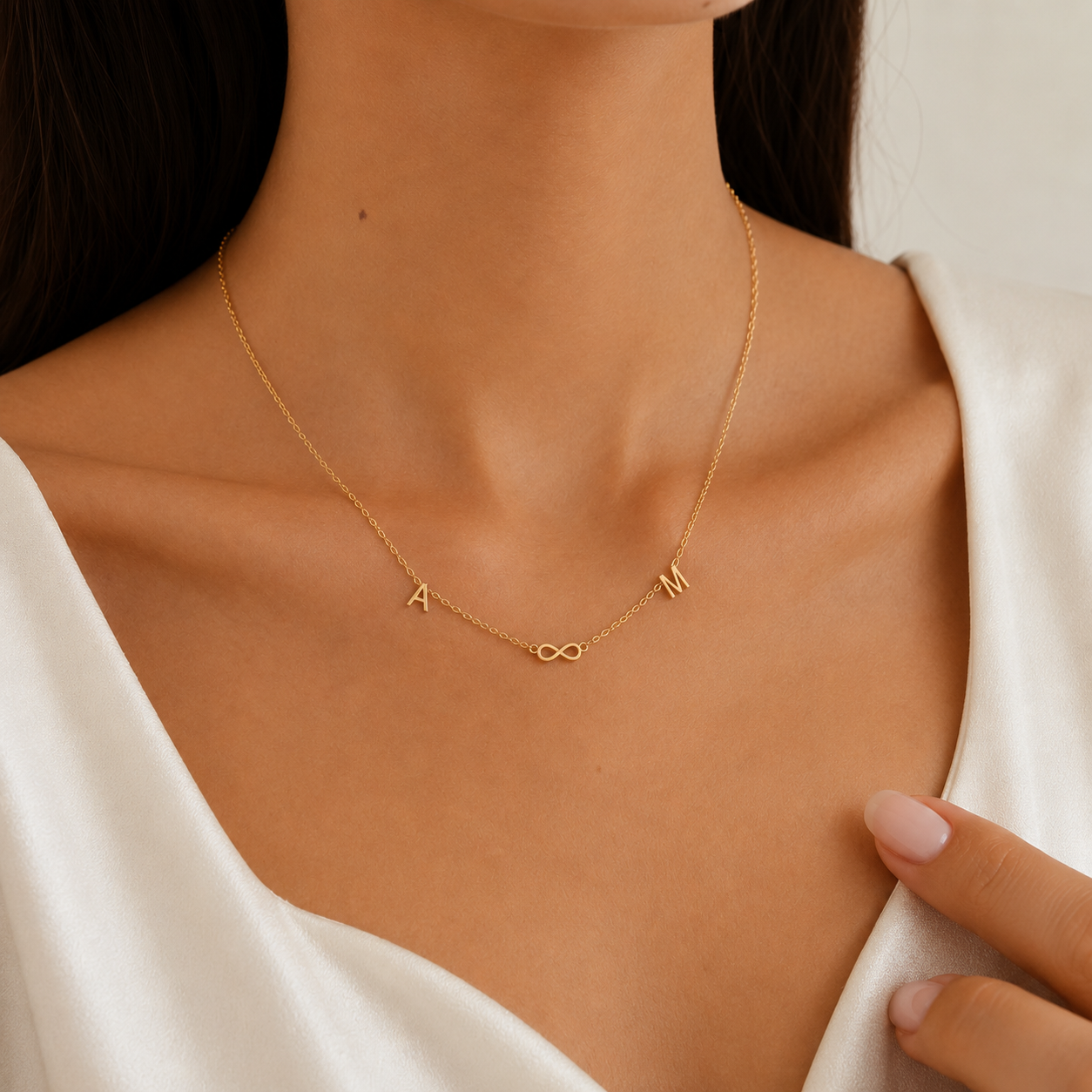 A close-up of a model wearing a delicate gold chain necklace with custom letter charms spelling 'A M' and an infinity symbol, styled with a white satin top.