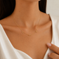 A close-up of a model wearing a delicate gold chain necklace with custom letter charms spelling 'A M' and an infinity symbol, styled with a white satin top.