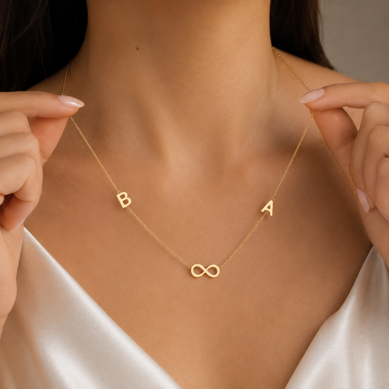 A delicate gold necklace worn by a model, featuring the letters 'B' and 'A' on either side of an infinity symbol at the center. The model is wearing a white satin top, and her hands are gently holding the chain to show its flexibility.