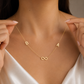 A delicate gold necklace worn by a model, featuring the letters 'B' and 'A' on either side of an infinity symbol at the center. The model is wearing a white satin top, and her hands are gently holding the chain to show its flexibility.