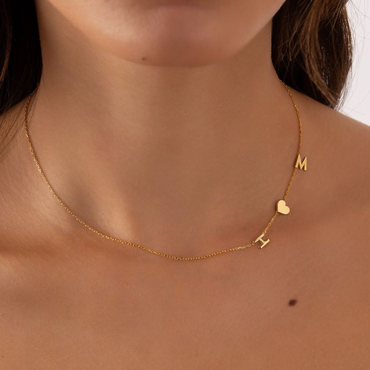 A delicate gold chain necklace worn by a model, featuring small letter charms 'M' and 'H' and a heart charm.