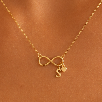 A delicate gold necklace with an infinity symbol pendant, a small heart charm, and a dangling letter 'S', displayed on a warm brown skin background.