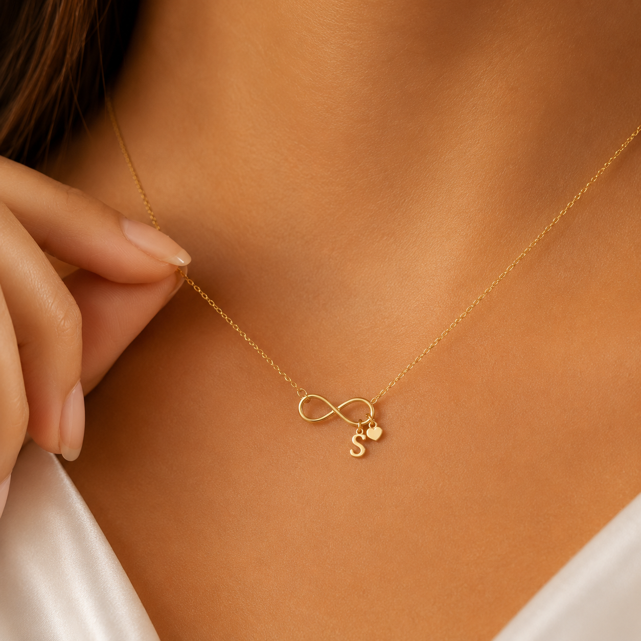 A delicate gold necklace worn on a model's neck, featuring an infinity symbol pendant with a small heart and the letter 'S' charm. The model is wearing a white satin top, and her hand gently touches the chain.