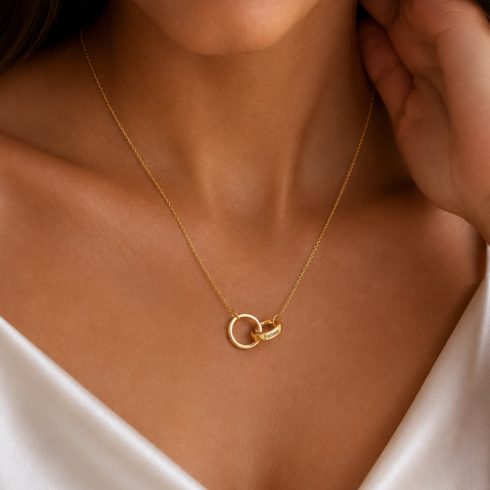 A close-up of a woman wearing a delicate gold necklace with two interlocking rings pendant, styled with a white satin camisole.