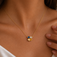 A close-up lifestyle shot of a model wearing a delicate gold chain necklace with a small four-leaf clover charm. The charm features one blue heart, one clear heart, and one gold heart.