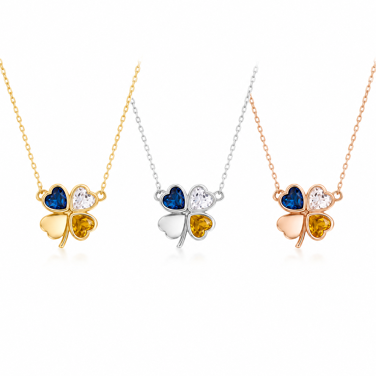 Three four-leaf clover pendant necklaces on delicate chains, shown in gold, silver, and rose gold finishes. The center clover features a blue heart, a clear heart, and a yellow heart, while the side clovers feature matching metal finishes with clear and yellow hearts.