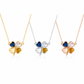 Three four-leaf clover pendant necklaces on delicate chains, shown in gold, silver, and rose gold finishes. The center clover features a blue heart, a clear heart, and a yellow heart, while the side clovers feature matching metal finishes with clear and yellow hearts.