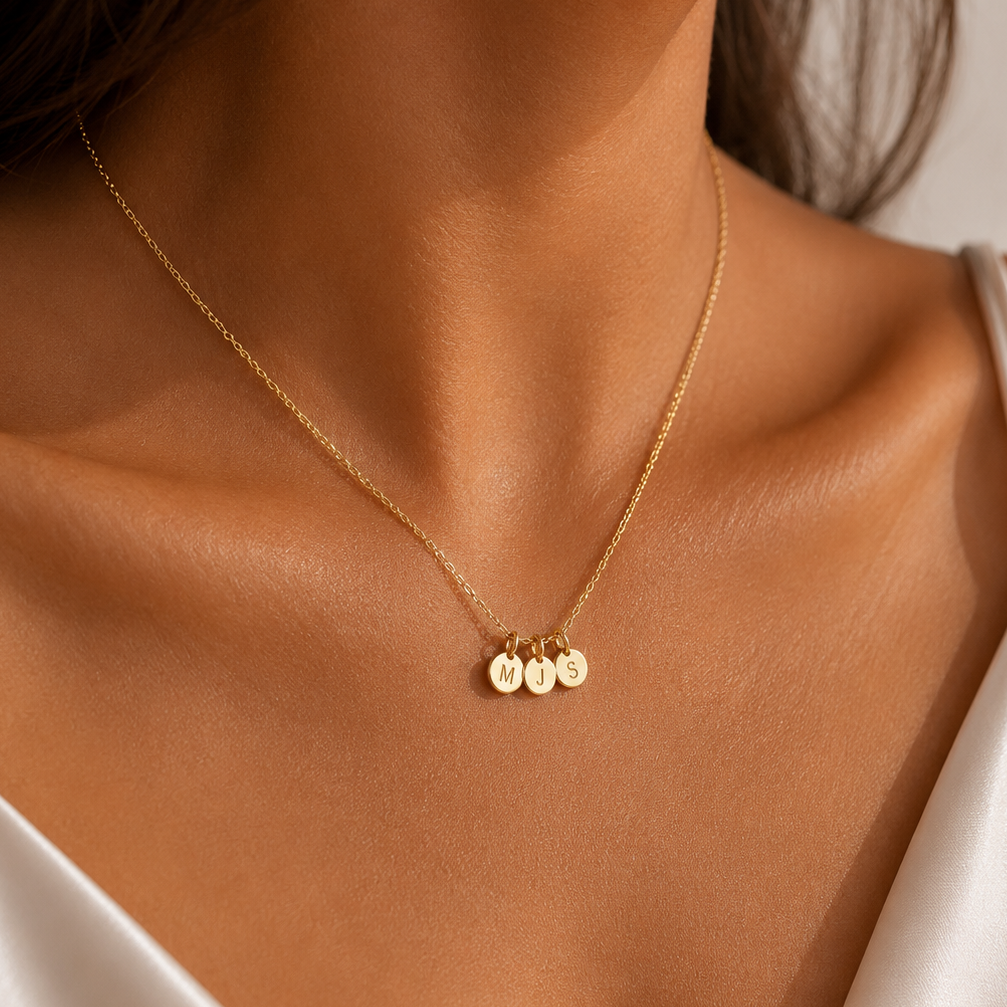 A close-up of a model wearing a delicate gold chain necklace with three small round engraved nameplates, styled with a white satin camisole.