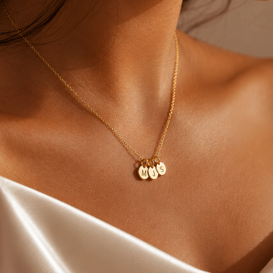 A close-up lifestyle shot of a woman wearing a delicate gold chain necklace with three small round gold pendants engraved with letters, styled with a white satin top.