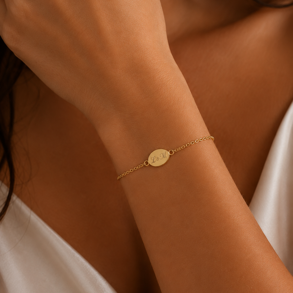 A delicate gold chain bracelet with a small oval engraved charm worn on a woman's wrist, shown in a close-up lifestyle shot.