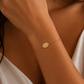A delicate gold chain bracelet with a small oval engraved charm worn on a woman's wrist, shown in a close-up lifestyle shot.