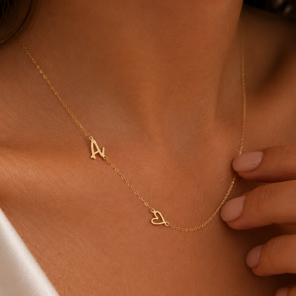 A close-up of a model wearing a delicate gold chain necklace with two small initial pendants, one shaped like the letter 'A' and the other like a heart containing a small heart against a warm skin tone.