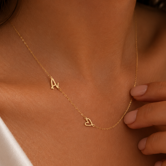 A close-up of a model wearing a delicate gold chain necklace with two small initial pendants, one shaped like the letter 'A' and the other like a heart containing a small heart against a warm skin tone.