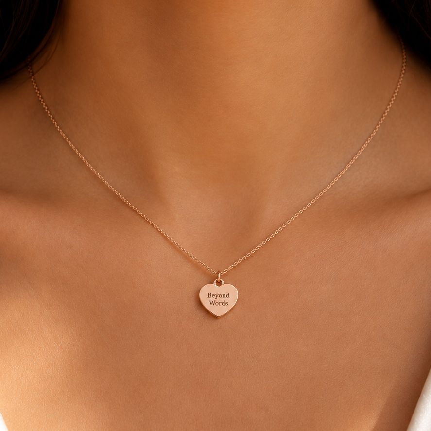 A close-up of a model's neck and chest wearing a delicate rose gold chain necklace with a small heart-shaped pendant engraved with 'beloved forever'.