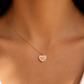A close-up of a model's neck and chest wearing a delicate rose gold chain necklace with a small heart-shaped pendant engraved with 'beloved forever'.